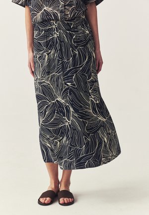Woman wearing a black midi skirt with large white floral outlines and black slide sandals against a plain background.