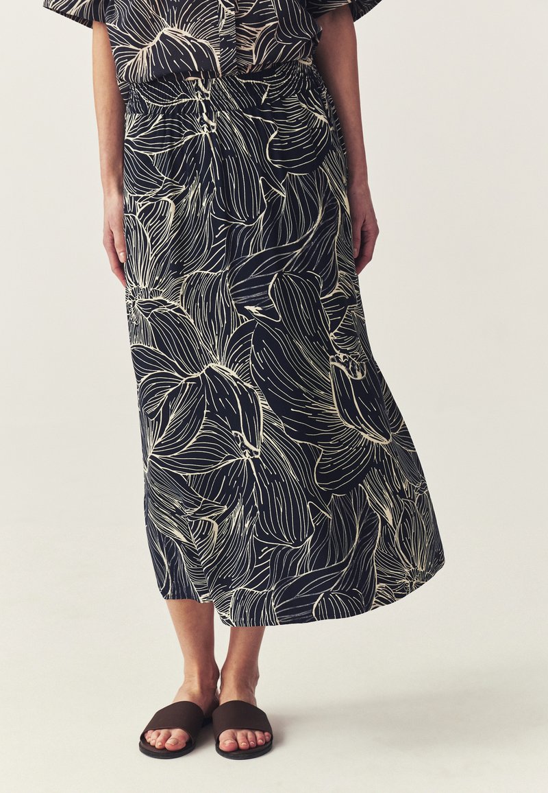 Femme portant une jupe midi noire avec de grands contours floraux blancs et des sandales noires à enfiler sur un fond uni.