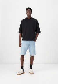 Μαύρο φαρδύ κοντομάνικο πουκάμισο, ανοιχτό τζιν σορτς, μαύρες κάλτσες και κρεμ αθλητικά παπούτσια με καφέ σόλες. Στηρίζει σε λευκό φόντο.