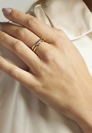 Goldring mit drei Bändern, zwei glatt und eines mit kleinen klaren Steinen, auf einer Hand mit heller Hautfarbe getragen. Weicher Stoff-Hintergrund.