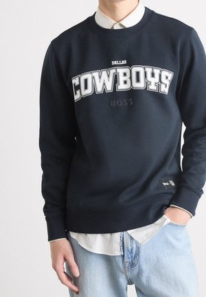 Person trägt ein dunkelblaues Sweatshirt mit den Logos "Dallas Cowboys" und "BOSS", dazu ein weißes Shirt und hellblaue Jeans.