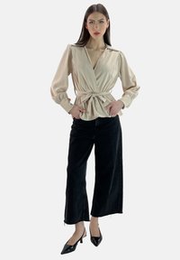 Blusa beige avvolgente con maniche lunghe e vita annodata, abbinata a pantaloni in denim neri a gamba larga e scarpe con tacco nere.