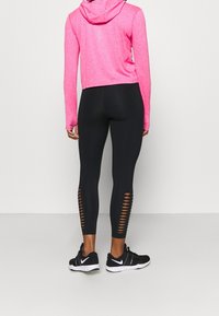 Roze cropped hoodie gecombineerd met zwarte leggings met uitsparingen aan de onderzijde van de benen en zwarte sportschoenen.