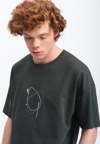 Donkergroene katoenen t-shirt met een eenvoudig abstract grafisch ontwerp in lichtblauw en paars, met een losse pasvorm en korte mouwen.