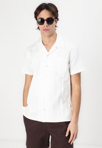 Camisa blanca de botones de manga corta con un patrón texturizado, combinada con pantalones cortos marrones. El modelo lleva gafas de sol negras.