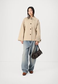 Beige oversized jakke med store brune knapper, kombinert med lyseblå vide bukser og en liten mørkebrun veske med skulderreim.