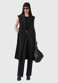 Donna che indossa un lungo cappotto senza maniche nero, top nero, jeans neri, stivali neri a punta, occhiali da sole, mentre tiene una grande borsa di pelle nera.