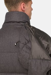 Vīrietis ar pelēku piepildītu jaku, kas rāda atvērtu daļu ar gludu oderi un regulējamu šņoru uz pleca.