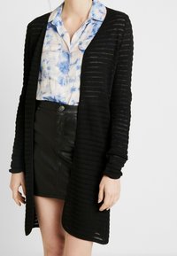 Černý pletený kardigan s otevřeným předním dílem, dlouhými rukávy a texturovaným pruhovaným vzorem. Nesený přes modré tričko s batikovaným vzorem a černou sukni.