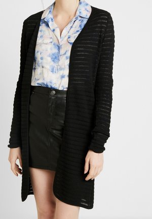 Cardigan en tricot noir à l'avant ouvert, à manches longues et à motif de rayures texturées. Porté par-dessus une chemise bleu tie-dye et une jupe noire.