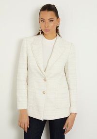 Blazer in tessuto avorio con colletto a punta, due tasche frontali e bottoni dorati, abbinato a un top crema e jeans blu scuro.