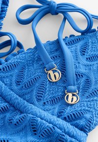 Blauw bikini-top gemaakt van textuurstof met bladerenpatronen, voorzien van gouden metalen accenten en verstelbare strikjes in een effen blauwe kleur.