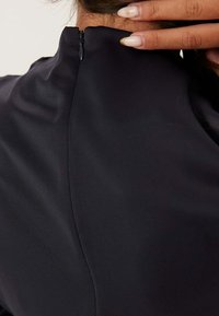 Svart klänning med hög halsringning och en dold dragkedja på baksidan; slätt, lätt material med en subtil lyster.