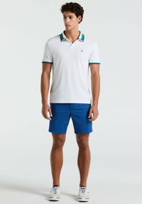 Weißes Polohemd mit kurzen Ärmeln und einem Kragen mit schwarzen, grünen und blauen Streifen sowie passenden Ärmelakzenten, kombiniert mit blauen Shorts und weißen Sneakers.