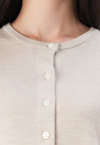 Beige strikkegenser med rund hals og fem hvite knapper foran. Stoffet har en glatt tekstur.