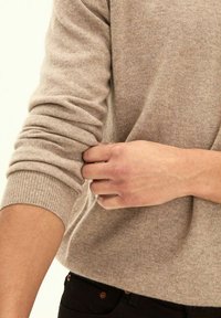 Helle beige Strickpullover mit gerippten Bündchen, der eine weiche Textur aufweist. Eine Hand zieht am Ärmel und zeigt ein einfaches, klassisches Design.