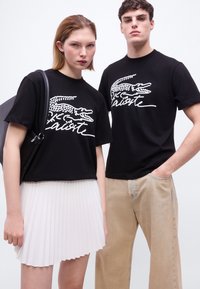 Δύο μοντέλα φοράνε μαύρα T-shirts με λευκό λογότυπο κροκοδείλου και επιγραφή· η γυναίκα φορά επίσης λευκή πλισέ φούστα και μαύρη τσάντα ώμου.