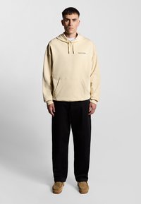Creme hoodie med snøre og frontlomme, parret med sorte bukser og brune sko. Hoodien har et lille logo på brystet.