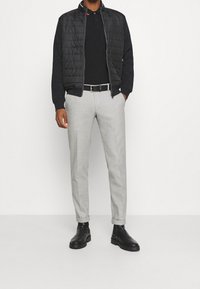 Zwarte gewatteerde jas, zwart poloshirt, lichtgrijze op maat gemaakte broek en zwarte enkellaarsjes met gestructureerde zolen. Geen zichtbare patronen of accenten.