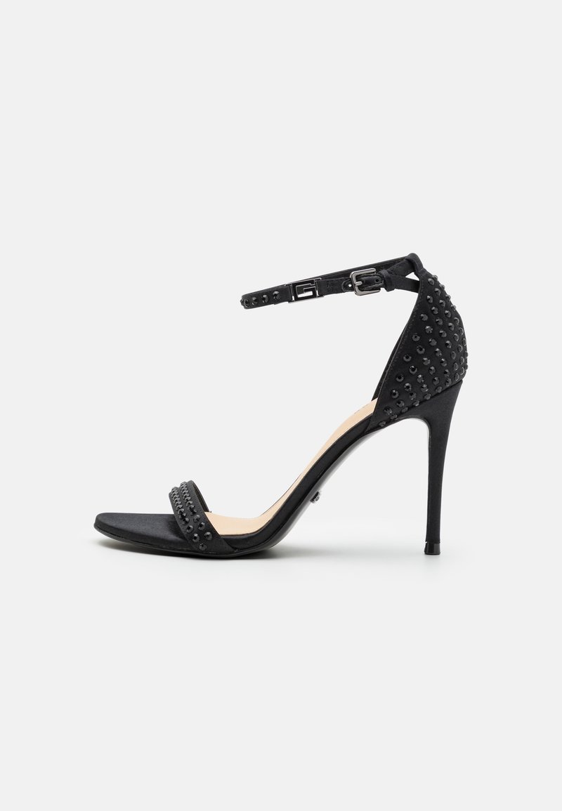 Sandale noire à talon haut avec bride à la cheville, bout ouvert et détails cloutés sur le talon et les brides, vue de profil sur fond blanc.