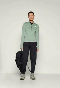 Lysegrønn genser med glidelås, svarte bukser, grå sko. Personen holder en svart ryggsekk; logo synlig på genseren og buksene. Glatt stoff, avslappet passform.