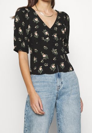 Femme portant un chemisier noir à manches courtes avec un imprimé floral rose et des boutons, associé à un jean taille haute bleu clair et un collier en or.