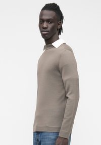 Jasnobrązowy dziany sweter o fakturowanej powierzchni, z białym kołnierzem i długimi rękawami, noszony z niebieskimi dżinsami na jednolitym tle.