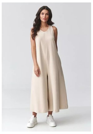 Kobieta z długimi falującymi włosami, ubrana w bezrękawnikowy, beżowy kombinezon z szerokimi nogawkami i białe sneakersy na platformie, stojąca na tle jasnego, jednolitego tła.