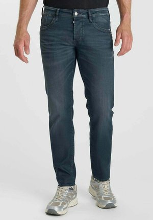 Mann trägt dunkelblaue Slim-Fit-Jeans und graue Sportschuhe und steht vor einem einfarbigen Hintergrund.