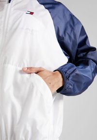 Giacca a vento bianca e blu navy con tasca frontale, polsini elasticizzati e logo sul petto. Tessuto liscio e leggero.