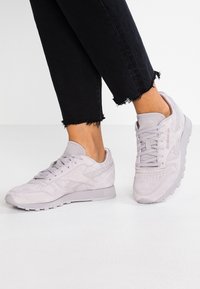 Ljust grå mockaskor med texturerade accenter, rund tå, snörning och logodetalj, kombinerat med svarta jeans med fransar.