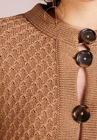 Cardigan lavorato a maglia in una calda tonalità di marrone, caratterizzato da un motivo testurizzato, un colletto rotondo e grandi bottoni scuri all'apertura frontale.