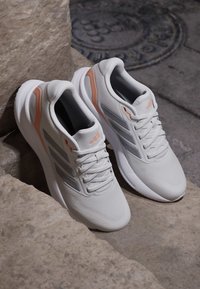 Chaussures de sport blanches avec des accents pêche reposant sur des marches en pierre près d'une surface en béton texturée et d'une plaque de regard en métal.