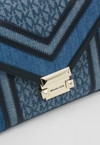 Pochette en denim présentant un motif chevron dans des tons bleus, avec des accents noirs et un fermoir doré orné de clous pyramidaux.