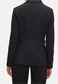 Personne portant un blazer noir ajusté et un pantalon noir assorti, vue de dos sur un fond gris uni.