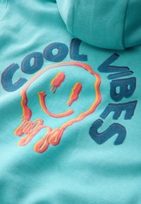 Türkisfarbener Hoodie mit einem grafischen Design, das die Worte "COOL VIBES" und ein tropfendes Smiley-Gesicht in lebendigen Farben zeigt. Weiche Stoffstruktur.