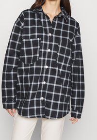 Kvinna klädd i en lös svart- och vitrutig skjorta med knappar och två bröstfickor, kombinerad med ljusbeiga byxor.