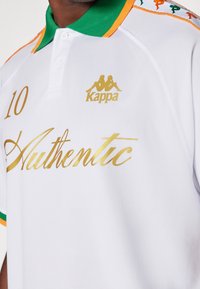 Bílá polokošile s zelenými a oranžovými detaily. Vyznačuje se zlatým nápisem "10" a "Authentic", s logem Kappa nahoře. Texturovaná tkanina.