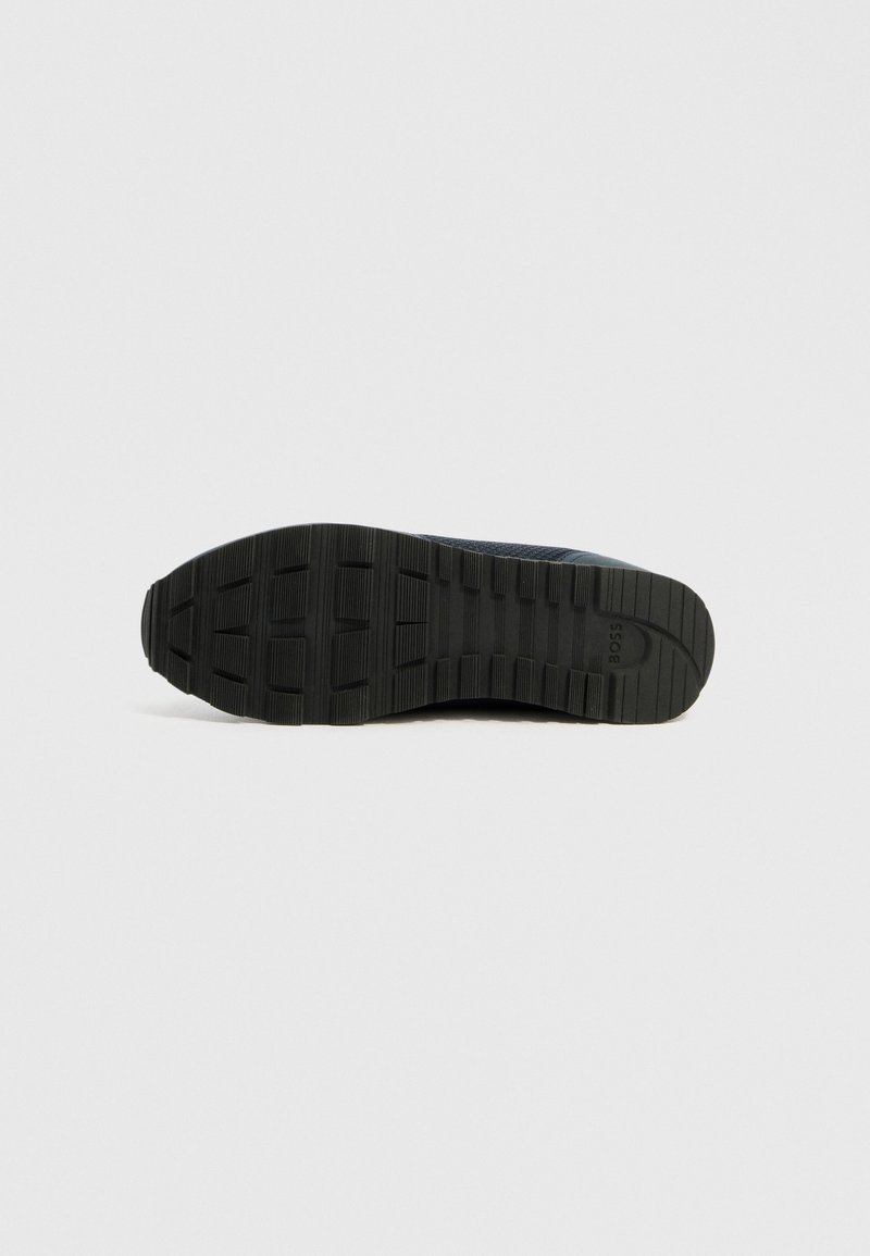 Semelle de sneaker noire avec une surface texturée et des crampons profonds pour une bonne adhérence. Comprend un bout arrondi et un logo discret sur le côté.