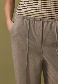 Pantaloni maro, texturati, cu un luciu discret, având închidere cu nasture, buzunare laterale și o croială lejeră. Parte dintr-un top cu dungi este vizibilă deasupra.