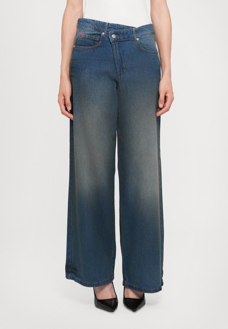 Personne portant un jean bleu délavé, à jambes larges et oversized, avec des talons noirs pointus et un haut blanc, debout devant un fond uni.