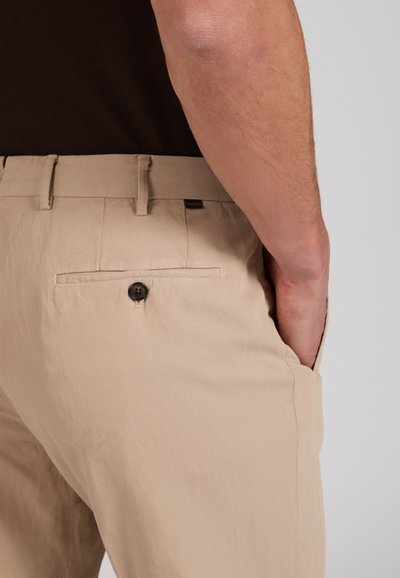 Pantalon beige avec poche arrière boutonnée porté par une personne avec la main dans la poche latérale, portant un haut foncé, fond clair.