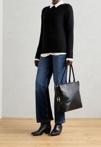 Personne portant un pull noir par-dessus une chemise blanche à col, un jean bleu à jambes larges, des bottines noires et tenant un grand sac cabas en cuir noir.