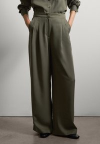 Personne portant un pantalon large vert olive et un haut à manches longues assorti, les mains dans les poches, sur un fond uni.