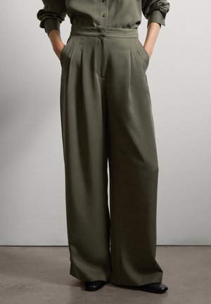 Persoon in olijfgroene wijde broek en bijpassend langemouwen top met handen in de zakken tegen een effen achtergrond.