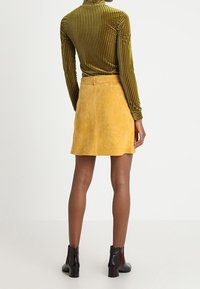 Jupe en daim jaune à silhouette droite, associée à un haut vert côtelé à manches longues et des bottines noires à finition brillante.