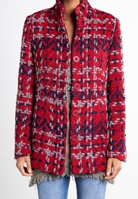 Persona che indossa una giacca di lana a trama rossa, blu navy e grigia con quattro bottoni, sopra un top grigio con frange e jeans blu.