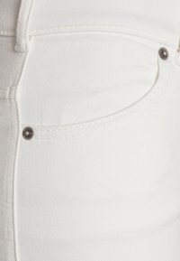 Close-up de jeans de ganga brancos com um bolso frontal, costura visível e detalhe em botão de metal. A textura parece lisa e ligeiramente texturizada.