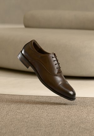 Chaussure habillée en cuir marron avec lacets flottant au-dessus d'un tapis marron clair, fond neutre avec canapé flou.