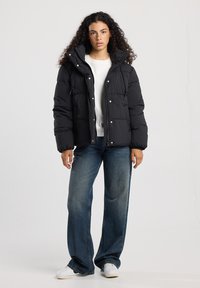 Doudoune noire avec capuche, boutons-pression et cordons de serrage. Portée sur une chemise blanche et assortie à un jean large bleu clair et des baskets blanches.